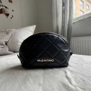 Svart quiltad handväska Valentino - Snygg svart handväska från Mario Valentino med quiltad design och guldfärgad dragkedja. Väskan har rundad form, diskret logga framtill och innerfack med dragkedja. Perfekt storlek för det viktigaste och ger en lyxig känsla till din outfit.