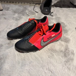 Nike Phantom Vision fotbollsskor svart/röd - Säljer ett par Nike Phantom Vision fotbollsskor i svart och rött med silverfärgad swoosh. Skorna har snörning, texturerad ovandel för bättre bollkontroll och en smal, sportig siluett. Perfekta för dig som vill sticka ut på planen. Namnen på skorna är borta