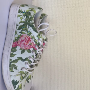 Blommiga vita sneakers med snörning - Snygga vita sneakers med färgglatt blommönster i grönt och rosa. Skorna har klassisk rund tå, vita snören och platt sula. Perfekta för dig som vill sticka ut med en fräsch och somrig outfit . De är riktigt lätta att matcha till olika outfits.