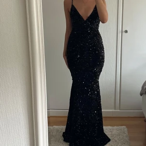 Svart glittrig långklänning Faeriesty - Supersnygg svart långklänning från Faeriesty med smala axelband, djup v-ringning och figurnära passform. Klänningen är täckt av glittrande paljetter och har öppen rygg, perfekt för att sticka ut på bal eller fest.