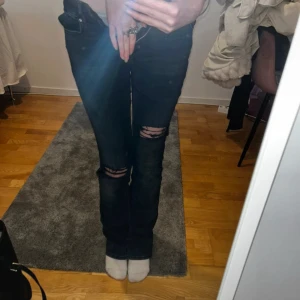Mörkblå slitna bootcut jeans - Säljer ett par mörkblå jeans med slitningar över knäna. Modellen har bootcut-ben och klassisk femficksdesign. Jeansen är i stretchigt denimtyg och har låg midja. Perfekta för en avslappnad och trendig look.
