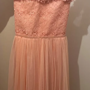 Ljusrosa långklänning med spets och pärlor, stl 152 - Romantisk ljusrosa långklänning med blommig spets upptill, pärldetaljer i midjan och volangprydd axel. Kjolen är i tunt, svepande tyg och överdelen har smockad rygg för skön passform. Perfekt för bal eller student!