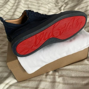 Mörkblå mocka sneakers från Louboutin - Äkta & Snygga mörkblå sneakers i mocka från Christian Louboutin med ikonisk röd sula och diskret logga på hälen. Skorna har rund tå, platt sula och klassisk snörning. Perfekta för dig som vill sticka ut med exklusiv stil och lyxiga detaljer. Kvitto finns