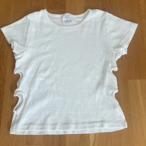 Vit ribbad topp med cutouts från Zara - Säljer en vit ribbad topp från Zara i storlek 164. T-shirten har korta ärmar och coola vågformade cutouts längs sidorna som ger en unik look. Perfekt för dig som vill sticka ut lite extra! Materialet är mjukt och stretchigt för en bekväm passform.