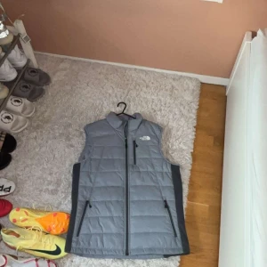 Grå dunväst från The North Face - Snygg grå dunväst från The North Face med quiltad design och svarta detaljer längs sidorna. Västen har hög krage, tre dragkedjefickor och hel dragkedja framtill. Perfekt att slänga över en hoodie för extra värme.