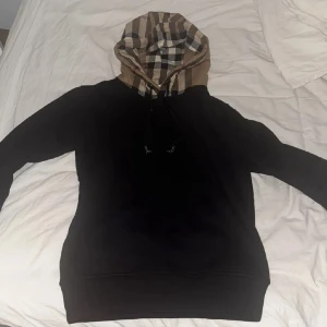 Svart Burberry hoodie med rutig huva - Här har vi en riktigt go burberry hoodie med den klassiska rutiga luvan, perfekt till både lite finare tillfällen men även en klassisk vardag.