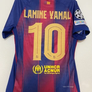 FC Barcelona Lamine Yamal matchtröja - Snygg FC Barcelona matchtröja med Lamine Yamal och nummer 10 på ryggen. Tröjan är kortärmad, har klassiska blå och röda ränder, gula detaljer och klubbmärke på bröstet. Materialet är lätt och andas, perfekt för fotboll eller att bära som supporter.