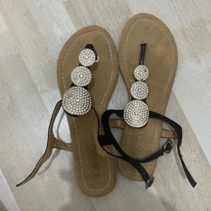 Svarta sandaler med strassdetaljer - Snygga svarta sandaler från Dona Girl med tre stora runda strassprydda dekorationer på ovansidan. Remmar i svart material med spänne vid ankeln och platt sula i beige ton. Perfekta för att ge lite extra bling till din sommaroutfit.