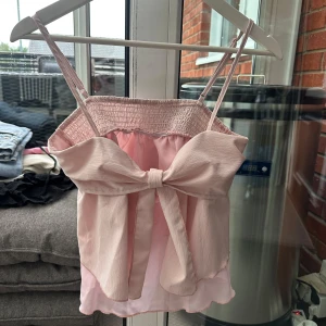 Ljusrosa linne/topp med rosett S - Supersöt ljusrosa linne/topp med smala reglerbara  axelband och stor rosett framtill. Toppen har smockad överdel och lätt volangig nederkant. Storlek S 