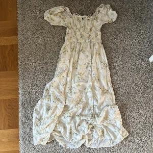 Blommig långklänning från Hollister - Supersöt långklänning från Hollister i ljus, nästan vit ton med blommigt mönster i gult och grönt. Klänningen har puffiga korta ärmar, smockad midja och knytning vid halsen. Perfekt för sommarens alla tillfällen!