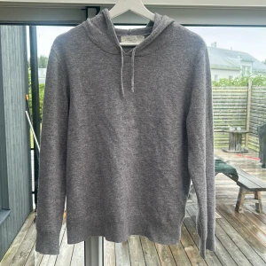 Grå kashmirhoodie  - Mjuk och lyxig grå hoodie i 100% kashmir. Tröjan har huva med snörning och långa ärmar, perfekt för dig som gillar en clean och stilren look. Ribbstickade muddar vid ärmslut och nederkant ger en snygg finish.