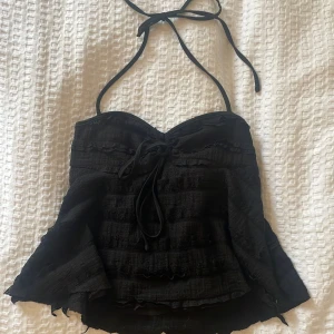 Svart halterneck topp med volanger - Supersnygg svart halterneck topp med volangdetaljer och snörning framtill. Toppen har en lätt utsvängd form och är tillverkad i ett strukturerat tyg som ger en cool look. Perfekt för dig som vill sticka ut med en trendig och edgy vibe.