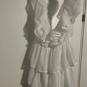 Vit volangklänning från NLY Eve - Superfin vit kortklänning från NLY Eve med flera lager volanger och djup v-ringning. Klänningen har en luftig känsla och är perfekt för sommarens alla tillfällen. Snygg omlott-look upptill och markerad midja för en feminin siluett.