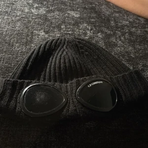 Svart C.P. Company goggle-mössa - Säljer en svart ribbstickad mössa från C.P. Company med ikoniska goggle-linser framtill. Mössan är tillverkad i mjukt material och har en tight passform som sitter snyggt på huvudet. Perfekt för dig som vill sticka ut med streetwear-vibe.