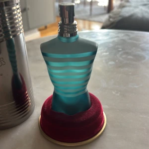 Jean Paul Gaultier Le Male parfym - Ikonisk parfymflaska formad som en manlig torso i frostat turkost glas med ljusblå ränder. Kommer med original metallburk och står på en röd sammetsliknande bas. En klassiker med unik design som sticker ut i badrumshyllan. Ordinarie pris cirka 800kr. 
