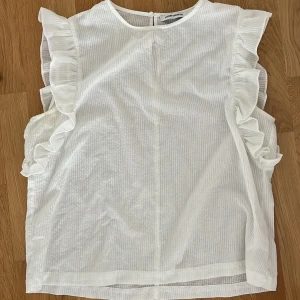 Vit blus med volang från Carin Wester - Supersöt vit blus från Carin Wester med diskret randig struktur och volangdetaljer längs ärmhålen. Blusen är ärmlös och har en liten knapp i nacken. Perfekt för dig som gillar en romantisk och stilren look.🌸