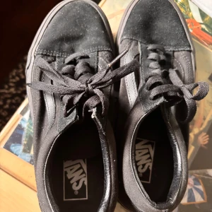 Svarta Vans Old Skool sneakers - Klassiska svarta Vans Old Skool sneakers med svart sula och ikonisk sidorand. Ovandel i canvas och detaljer i skinn, med snörning och den typiska gummisulan i brunt. Perfekta för en avslappnad streetstil.