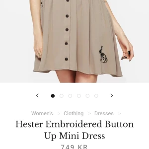 Beige broderad kortklänning med knappar - Supersöt beige kortklänning med svarta broderier, v-ringning och knappar hela vägen framtill. Klänningen har korta ärmar och coolt broderat skelett på kjolen samt blommor på axlar och rygg. Perfekt för dig som gillar unika detaljer och chill vibe. Skickas ✨