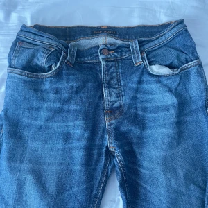 Nudie Jeans blå Slim Tim jeans 32/32 - Säljer ett par klassiska Slim Tim jeans från Nudie Jeans i mellanblå tvätt. Jeansen har orangea sömmar, fem fickor och snygga detaljer på bakfickorna. Tillverkade i bomull med en rak och något smalare passform. Perfekta för dig som gillar stilrena jeans med cool edge.