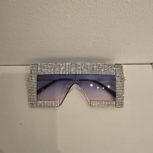 Glittriga statement-solglasögon - Riktigt feta solglasögon med stora, rektangulära linser i lila ton och en ram täckt av glittrande stenar. Perfekt för dig som vill sticka ut och maxa din look. Linsen är tonad och ramen är kantig för en futuristisk vibe.