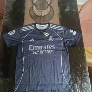 Real Madrid tröja  - Snygg Real Madrid fotbollströja från Adidas i mörkblått med vita detaljer. Tröjan har Mbappé och nummer 9 på ryggen, klubbmärke på bröstet och Emirates Fly Better-tryck. Tillverkad i lätt och ventilerande material, perfekt för match eller träning.