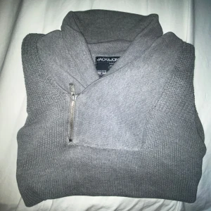 Grå stickad tröja från Jack & Jones - Säljer en grå stickad tröja från Jack & Jones med hög krage och snygg half zip-dragkedja. Tröjan har långärmade ärmar och olika stickade strukturer på axlar och muddar. Perfekt för dig som gillar stilren och enkel design.