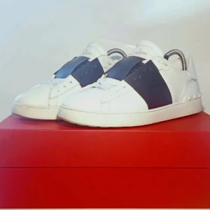 Valentino Open sneakers vit/blå - Snygga Valentino Open sneakers i vitt skinn med bred blå rem över vristen och ikoniska nitar baktill. Klassisk rund tå och platt sula ger en clean och modern look. Perfekta för dig som vill sticka ut med stilrena detaljer.