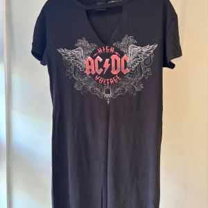 Svart AC/DC t-shirtklänning FB Sister - Svart t-shirtklänning från FB Sister med AC/DC High Voltage-tryck i rött och vitt framtill. Klänningen har korta ärmar, v-ringning med cool cutout och är gjord i mjuk bomull. Perfekt för dig som gillar rockig stil och vill sticka ut.