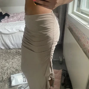 Beige långkjol med rynkade detaljer - Säljer en beige långkjol med snygga rynkade detaljer längs sidan och en liten slits nedtill. Kjolen har en tight passform och är gjord i ett mjukt, stretchigt material som sitter skönt på kroppen. Perfekt för dig som gillar stilrena och trendiga plagg.
