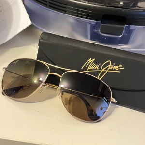 Guldiga aviator solglasögon Maui Jim - Snygga aviator solglasögon från Maui Jim med guldiga metallbågar och mörktonade linser. Klassisk pilotform som ger en cool vibe. Kommer med originalfodral och putsduk. Perfekta för soliga dagar och en stilren look. Sprillans nya kostar runt 3k kronor