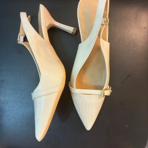 Beige pumps med spetsig tå och klack - Snygga beige pumps med spetsig tå och smal klack. Skorna har öppen häl med justerbart spänne i guld och en elegant rem över foten. Tillverkade i glansigt syntetmaterial som ger en stilren look. Aldrig använda endast testade där de var för små. 