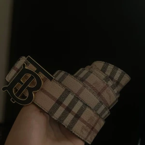 2 sidig b bälte  - Ett bälte med två sidor svart och beige med klassiskt rutigt mönster. Ena bältet är beige med svarta och röda rutor, det andra är svart med diskret rutmönster och präglad riddare. Båda har ett stort guldfärgat TB-spänne som ger en lyxig vibe. Tillverkade i skinn.