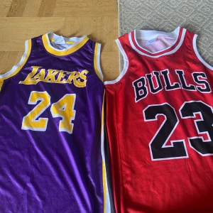 Lakers och Bulls baskettröjor - Två baskettröjor: en lila och gul Lakers-tröja med nummer 24 och en röd, vit och svart Bulls-tröja med nummer 23. Båda är ärmlösa och har klassiska klubbnamn tryckta på bröstet. Perfekt för basketfans eller samlare.