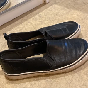 Svarta slip-on sneakers med vit sula  - Svarta slip-on sneakers med vit, platt sula och rund tå. Skorna har elastiska partier på sidorna för enkel på- och avtagning. Perfekta för en avslappnad och stilren look.