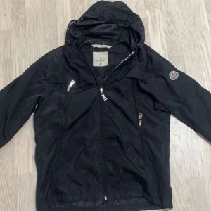 Svart vindjacka från Moncler med huva - Säljer en svart Moncler vindjacka med huva och dragkedja. Jackan har Moncler-logga på ärmen och tryckt Moncler-text på insidan av huvan. Flera fickor med dragkedja och lätt material som passar perfekt till vår och höst.