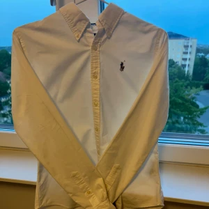 Ralph Lauren skjorta - Klassisk vit skjorta från Ralph Lauren med broderad logga på bröstet. Skjortan har lång ärm, knappar hela vägen och button-down krage. Tillverkad i bomull för en fräsch och stilren look. Perfekt för dig som gillar tidlös och clean.