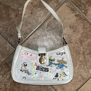 Vit axelväska med Toy Story-tryck - Vit axelväska i syntet med färgglatt Toy Story-inspirerat tryck framtill. Väskan har justerbart axelband, guldfärgat lås och lekfulla motiv av Buzz Lightyear, utomjordingar och texten LOVE. Perfekt för dig som gillar unika accessoarer med nördiga detaljer.