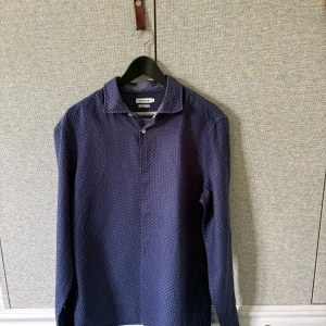 Mörkblå mönstrad skjorta från J.Lindeberg - Snygg mörkblå skjorta från J.Lindeberg med diskret prickigt mönster. Slim fit-modell med klassisk krage och knäppning framtill. Perfekt för dig som vill ha en stilren look med lite extra detalj. Men också till sånna som letar efter sommar skjorta eller en golf skjorta