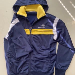  träningsjacka från Sergio Tacchini - Säljer en blå träningsjacka med gula och vita detaljer från Sergio Tacchini. Jackan har huva, dragkedja och ribbade muddar. Den är väldigt bra skick och den köptes ny pris för 2500kr den passar s och m
