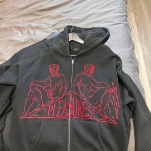 Svart hoodie med rött tryck från Pleasures - Svart hoodie från Pleasures med dragkedja och stort rött grafiskt tryck framtill. Trycket föreställer två figurer och texten 'PLEASURES'. Perfekt för dig som gillar streetwear och unika plagg.