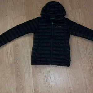 Svart dunjacka med huva från Peak Performance - Säljer en svart dunjacka från Peak Performance i storlek XS. Jackan har huva, dragkedja framtill och två sidofickor med dragkedja. Perfekt för kyliga dagar och enkel att matcha med allt.