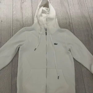 Vit hoodie från Lacoste med dragkedja - Snygg vit hoodie från Lacoste med klassisk krokodillogga på bröstet. Tröjan har huva med snörning, dragkedja framtill och två stora fickor. Perfekt för dig som gillar en clean och stilren look. Perfekt för sommaren! Helt ny, inga defekter alls. Pris kan diskuteras!