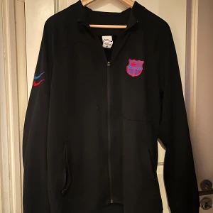 Svart Nike FC Barcelona zipper - Säljer en svart jacka från Nike med FC Barcelona-logga på bröstet och två broderade Nike-swooshes i blått och rött på ärmen. Jackan har dragkedja framtill och fickor med dragkedja. Texten 'CULERS' finns tryckt i nacken.