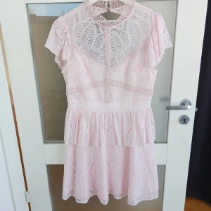 Ljusrosa spetsklänning med kort ärm - Säljer en söt ljusrosa klänning med spetsdetaljer och korta ärmar. Klänningen har hög hals, markerad midja och volang i kjolen. Perfekt för sommarens tillfällen eller studentfirande! Från By malina