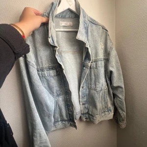 Ljusblå jeansjacka - Säljer en klassisk ljusblå jeansjacka med slitna detaljer och knappar framtill. Jackan har två bröstfickor och två sidofickor. Perfekt för dig som gillar en avslappnad och trendig stil.