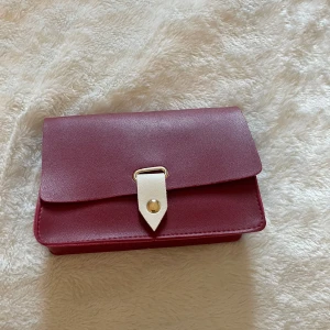 Vinröd axelväska med vit detalj - Säljer en stilren vinröd axelväska med guldfärgad knapp. Väskan har ett minimalistiskt och modernt utseende, perfekt för att bära det viktigaste. Materialet är slätt och väskan har en kompakt form.