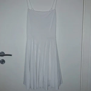 Vit kortklänning med tunna axelband - Superfin vit kortklänning med tunna axelband och snyggt korsad rygg. Klänningen har en lätt utsvängd kjol och enkel, stilren design som känns fräsch och somrig. Perfekt för dig som gillar minimalistisk stil och vill ha något luftigt och bekvämt.