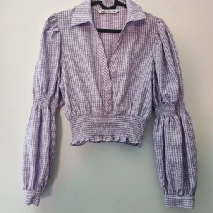 Lila rutig croppad blus från Zara - Säljer en lila och vit rutig croppad blus från Zara med långa puffärmar och smockad midja. Blusen har krage, v-ringning och är tillverkad i bomull. Perfekt för dig som gillar trendiga och feminina plagg!