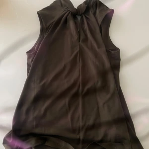 Svart ärmlös topp med knyt från H&M - Säljer en svart ärmlös topp från H&M med snygg knytdetalj vid halsen. Toppen har en rak passform och är tillverkad i ett mjukt material som känns skönt mot huden. Perfekt att styla med jeans eller kjol.