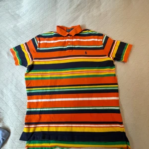 Randig pikétröja Polo Ralph Lauren L - Färgstark pikétröja från Polo Ralph Lauren i storlek L. Tröjan har breda ränder i orange, marinblått, grönt, gult och vitt, klassisk krage och knappar framtill. Tillverkad i 100% bomull och perfekt för dig som gillar en färgglad och chill stil.